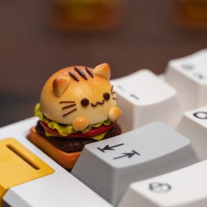 Könnte beinhalten: Eine skurrile, katzenförmige Burger-Tastenkappe. Die Tastenkappe hat ein Brötchen mit braunen Streifen, Katzenohren und einem lächelnden Gesicht. Der Burger enthält ein Patty, Salat und Tomate. Die Tastenkappe liegt auf einer weißen Tastatur.