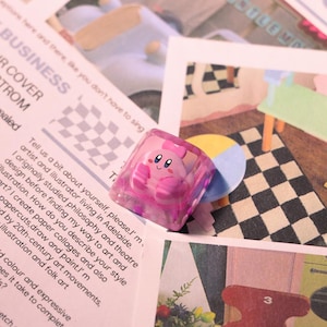 May include: A pink, translucent keycap featuring a cartoon character with a smiling face and a bow. The keycap is square-shaped and sits on a background of printed paper and illustrations. The keycap is a custom accessory for a mechanical keyboard.