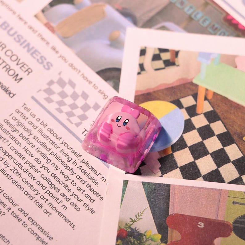 May include: A pink, translucent keycap featuring a cartoon character with a smiling face and a bow. The keycap is square-shaped and sits on a background of printed paper and illustrations. The keycap is a custom accessory for a mechanical keyboard.