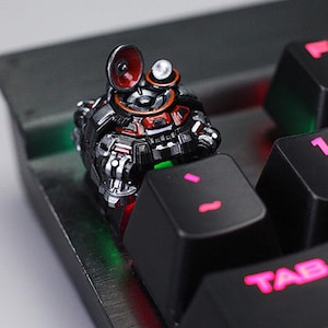 May include: A custom mechanical keyboard keycap featuring a detailed, futuristic robot design. The keycap is primarily black with red and silver accents, and a small speaker-like element. The keycap is on a black keyboard.