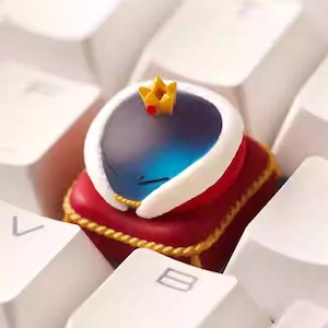 May include: A custom mechanical keyboard keycap shaped like a blue, translucent, rounded creature wearing a small yellow crown with a red accent, resting on a red square cushion base with white trim and gold rope detailing.