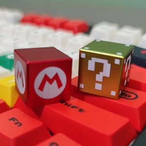 Könnte beinhalten: Zwei würfelförmige Tastenkappen mit Videospiel-Motiv. Eine ist rot mit weißem "M"-Logo, die andere goldfarben mit weißem Fragezeichen. Sie liegen auf einer roten Tastatur.