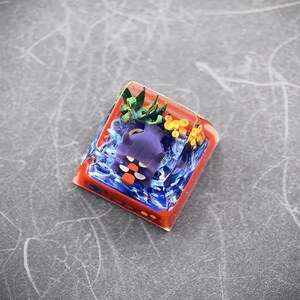 May include: A custom keycap featuring a purple creature with orange accents, set within a clear resin block. The design includes blue water effects, green foliage, and yellow floral elements, all encased in a red-orange frame.