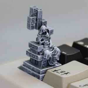 Puede incluir: Una tecla artesanal gris y detallada que representa una figura encapuchada sentada en una estructura similar a un trono. La tecla está diseñada para un teclado y tiene una apariencia desgastada, similar a la piedra. La tecla está en un teclado.