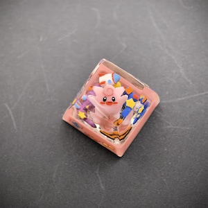 May include: A custom pink resin keycap with a cartoon character design. The keycap has a square shape and is decorated with blue, yellow, and purple accents. It is designed for use with a mechanical keyboard.