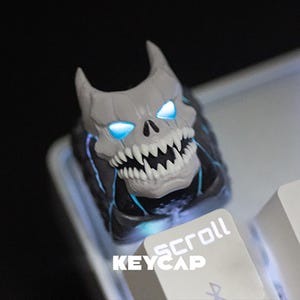 Può includere: Un copritasto a forma di teschio grigio con occhi blu luminosi e denti affilati. Il copritasto è su una tastiera bianca con la parola "KEYCAP" e "scroll" stampate sui tasti. Il teschio ha corna e un design dettagliato.