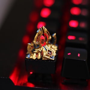 May include: A custom mechanical keyboard keycap with a detailed gold, blue, and red design. The keycap is mounted on a black base, with a red gem in the center. The background is a blurred view of a keyboard with red backlighting.