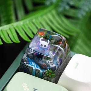 Könnte beinhalten: Eine individuelle Tastenkappe mit einer detaillierten Miniaturszene. Sie zeigt eine weiße Figur, blaues Wasser und eine Steinstruktur. Die Kappe ist transparent mit harzähnlichem Aussehen, vor grünem Hintergrund.