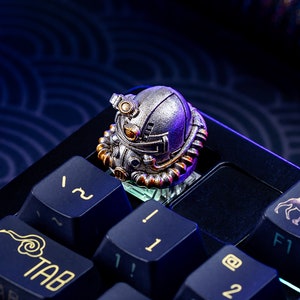 Peut inclure: Un keycap de clavier m&eacute;canique argent&eacute; et dor&eacute; en forme de casque futuriste. Le keycap est pos&eacute; sur un clavier bleu fonc&eacute; avec des lettres dor&eacute;es. Le keycap pr&eacute;sente des d&eacute;tails complexes et une finition m&eacute;tallique.