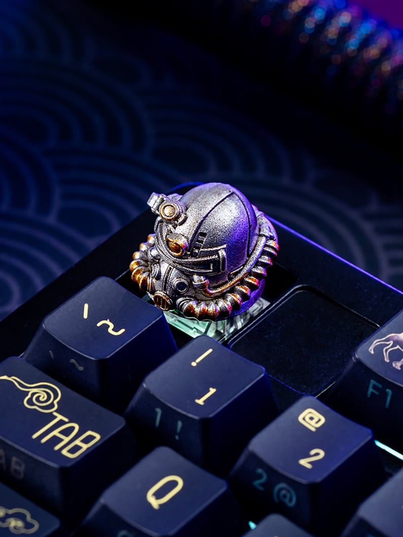 Peut inclure: Un keycap de clavier m&eacute;canique argent&eacute; et dor&eacute; en forme de casque futuriste. Le keycap est pos&eacute; sur un clavier bleu fonc&eacute; avec des lettres dor&eacute;es. Le keycap pr&eacute;sente des d&eacute;tails complexes et une finition m&eacute;tallique.