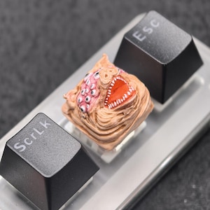 May include: A custom artisan keycap featuring a monster design with multiple eyes and an open mouth. The keycap is tan with pink and red accents. The keycap is flanked by black keyboard keys with "Esc" and "ScrLk" printed on them.