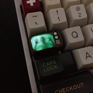 May include: A custom keyboard keycap featuring a miniature television design. The screen glows green, displaying a ghostly figure with outstretched hands. The keycap is surrounded by other white and black keys.