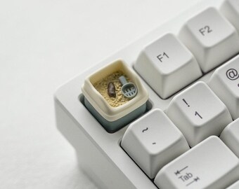 Puede incluir: Una tecla de teclado personalizada con un diseño de arenero para gatos en miniatura. La tecla es azul claro con un interior beige, con una pequeña pala y arena simulada. Las teclas circundantes son blancas.