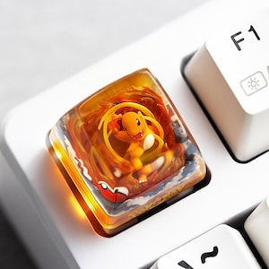 May include: A square artisan mechanical keyboard keycap featuring a translucent amber resin housing. Inside, a miniature orange Charmander figure is surrounded by swirling orange and yellow flame effects, with a small red and white ball detail.