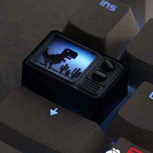 May include: A black custom keycap shaped like a retro television, featuring a pixelated dinosaur silhouette on a blue screen. The keycap has two round knobs and is set against a backdrop of gray keyboard keys.