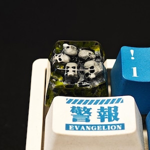 Puede incluir: Primer plano de una tecla de teclado personalizada con un diseño de varias pequeñas figuras de calaveras sobre una base verde y amarilla. La tecla de abajo tiene el texto "EVANGELION" en azul y blanco.