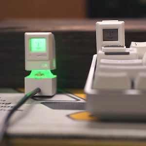May include: Two white miniature computer models with glowing green screens. One is plugged in with a green cable. The other is on a white keyboard. The screens display a smiling face. The overall theme is retro tech.