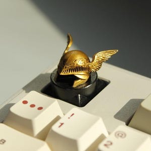 May include: A gold-colored helmet keycap with wing details, set on a black base, sits atop a white keyboard. The keycap has intricate detailing and is a decorative accessory for mechanical keyboards.