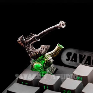 Könnte beinhalten: Ein detaillierter, Fantasy-Themen-Artisan-Keycap mit einem Schlachtbeil-Design. Der Keycap ist hauptsächlich braun und grün, mit aufwendigen Details und einem leuchtenden grünen Kern. Der Keycap befindet sich auf einer schwarzen Tastatur.