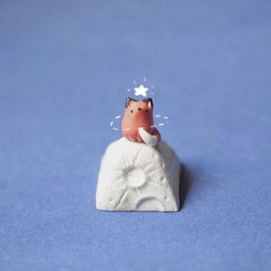 Puede incluir: Una pequeña figura de zorro hecha a mano en tonos coral, negro y blanco, posada sobre una base blanca texturizada en forma de luna. Una estrella blanca con un halo está encima del zorro. El fondo es azul liso.