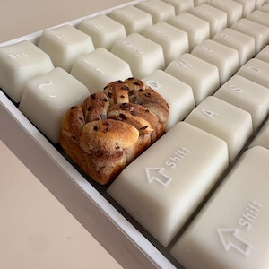 May include: A close-up shot of a keyboard with off-white keys. A loaf of bread with chocolate chips sits between the keys. The keyboard keys are arranged in rows and columns, with the letters and symbols visible.