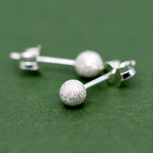 Peut inclure: Une paire de boucles d'oreilles en argent avec un motif de boule texturée. Les boucles d'oreilles sont sur un fond vert.