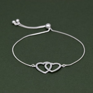 May include: A silver bracelet with two intertwined heart charms. The bracelet has a fine chain and a sliding bead clasp for adjustable sizing. The hearts are linked, representing unity and affection.