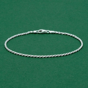 May include: A silver rope chain bracelet with a lobster clasp. The bracelet features a twisted, rope-like design and is set against a green backdrop. The bracelet is approximately 18 cm long.