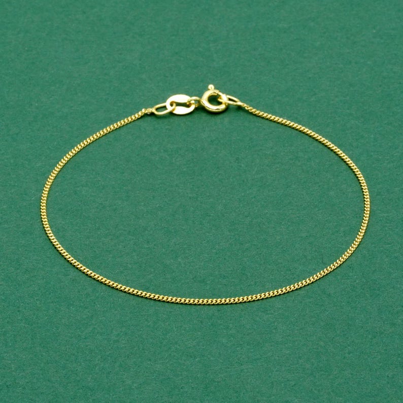 May include: A delicate gold-colored chain bracelet with a lobster clasp, set against a solid green background. The bracelet is made of small, interconnected links, creating a subtle, elegant design. The clasp is clearly visible.