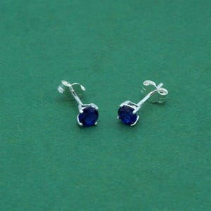 May include: A pair of stud earrings with a silver post and setting. Each earring features a round, deep blue gemstone. The earrings are set against a solid green background.