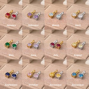 May include: A collection of twelve pairs of birthstone pendants, each featuring a round faceted gemstone in a gold or silver setting. The months January through December are labelled below each pair, showcasing colours like red, purple, blue, green, and yellow.