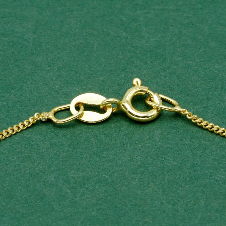 May include: Close-up of a gold-colored necklace clasp and chain. The clasp is a lobster claw style, connected to an oval-shaped tag. The chain has a delicate, curb link design. The background is a solid, dark green.