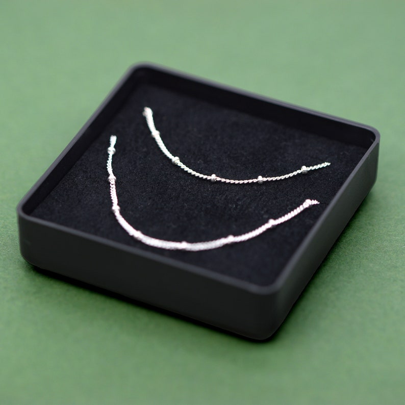 May include: Two silver necklaces with small beads, displayed in a black velvet-lined box.