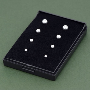 May include: A black jewellery box with a black velvet interior containing seven small, round, silver-coloured stud earrings. The earrings vary in size, with the smallest at the bottom and the largest at the top. The box is on a green surface.