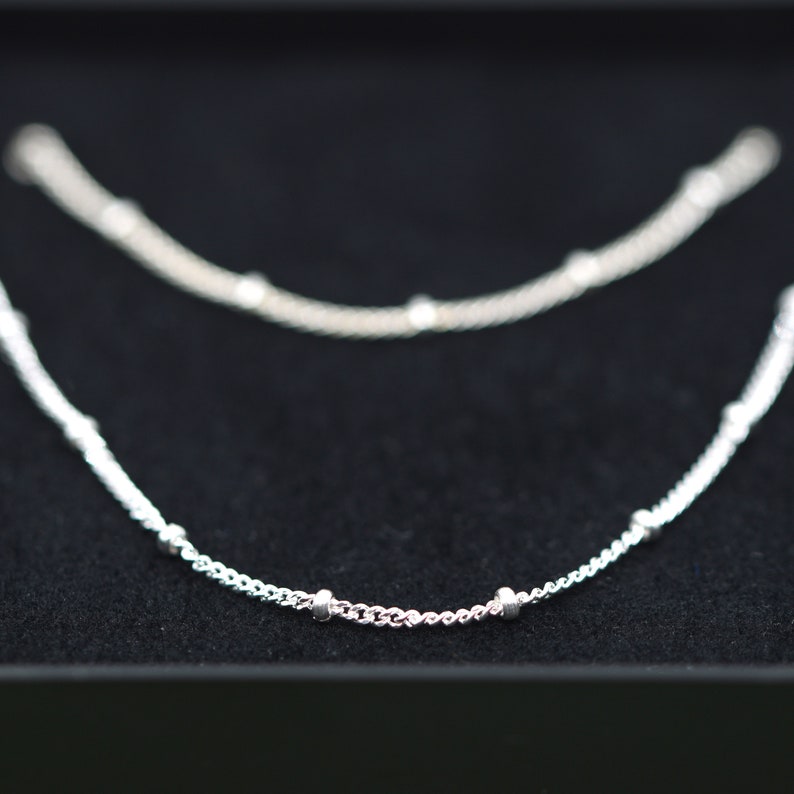 May include: Two silver necklaces with small round beads on a black background.