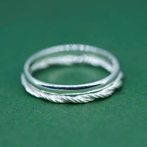 May include: A silver ring with a braided pattern around the band, set against a green backdrop. The ring has a polished surface, reflecting light. The design gives the ring a distinctive and elegant appearance.
