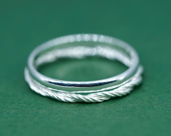 May include: A silver ring with a braided pattern around the band, set against a green backdrop. The ring has a polished surface, reflecting light. The design gives the ring a distinctive and elegant appearance.