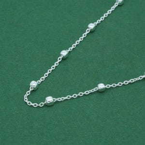 May include: A delicate silver necklace with a chain design. The necklace features small, square-shaped beads evenly spaced along the chain. The jewellery is displayed against a solid green background.