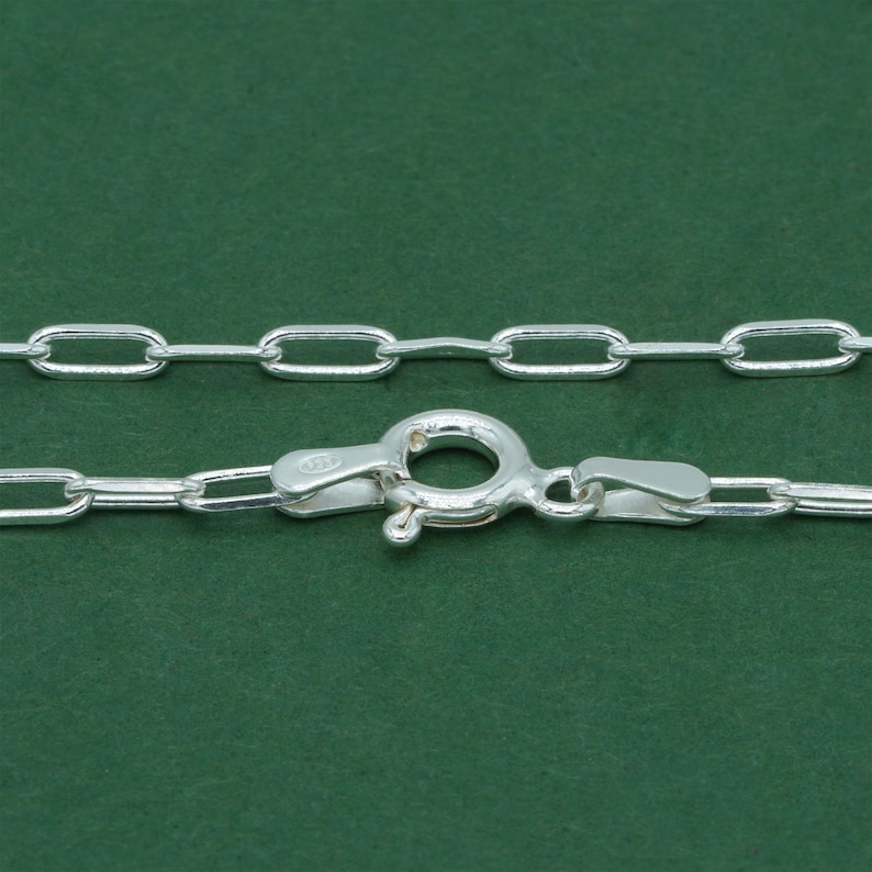 May include: Close-up of a silver chain with an oval link design. The chain features a lobster clasp for secure fastening. The links are bright and reflective against a green background.