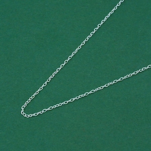 Könnte beinhalten: Eine zarte Silberkette vor grünem Hintergrund. Die Kette besteht aus kleinen, miteinander verbundenen Gliedern, die ein subtiles, elegantes Design ergeben. Die Halskette ist ideal zum Kombinieren oder allein tragen.