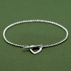 May include: A silver bracelet with a heart-shaped clasp. The bracelet is made of small, round beads.