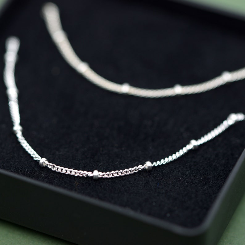 May include: Two silver chains with small round beads on a black velvet background. The chains are delicate and have a simple design.