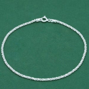 May include: A silver bracelet with a textured, sparkling design. The bracelet has a secure clasp and is shown against a green background. This piece of jewellery is suitable for everyday wear or special occasions.