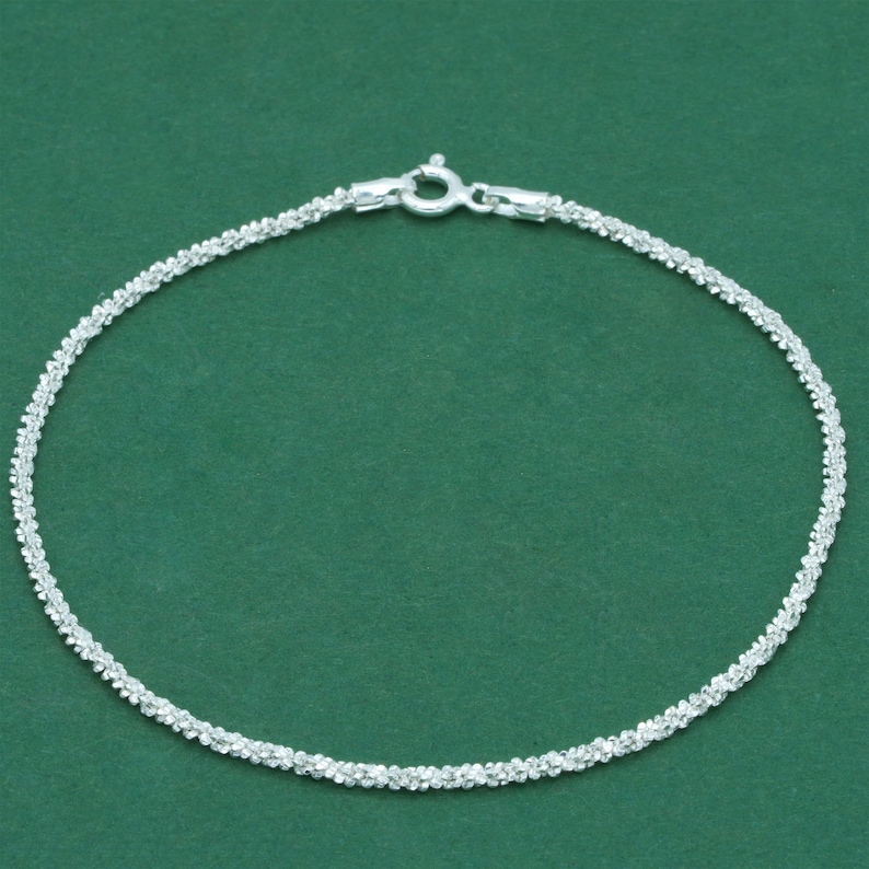 K&ouml;nnte beinhalten: Ein zartes Silberarmband mit strukturiertem, funkelndem Design. Das Armband verf&uuml;gt &uuml;ber einen sicheren Verschluss und wird vor einem einfarbigen gr&uuml;nen Hintergrund pr&auml;sentiert. Dieses Schmuckst&uuml;ck eignet sich f&uuml;r den Alltag oder besondere Anl&auml;sse.