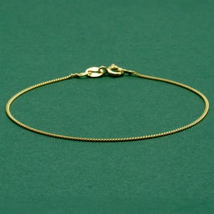 May include: A delicate gold-colored chain bracelet with a lobster clasp closure. The bracelet is displayed on a solid green background. The chain has a fine, textured design, and the clasp is a matching gold tone.