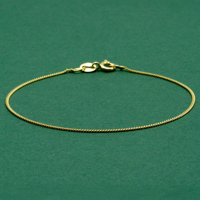 May include: A delicate gold-colored chain bracelet with a lobster clasp closure. The bracelet is displayed on a solid green background. The chain has a fine, textured design, and the clasp is a matching gold tone.
