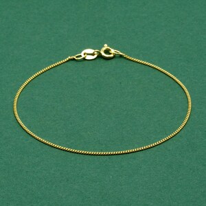 May include: A delicate gold-colored chain bracelet with a lobster clasp closure. The bracelet is made of a fine, textured chain, and is displayed against a solid green background. The bracelet is a simple, elegant piece of jewelry.