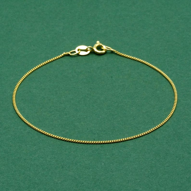 May include: A delicate gold-colored chain bracelet with a lobster clasp closure. The bracelet is made of a fine, textured chain, and is displayed against a solid green background. The bracelet is a simple, elegant piece of jewelry.