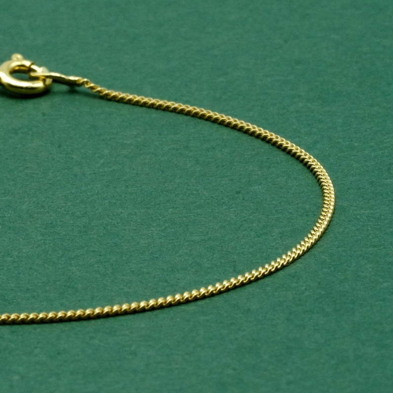 May include: A gold-colored chain necklace with a lobster clasp, set against a dark green background. The chain has a delicate, textured design, and the clasp is visible at the top left.