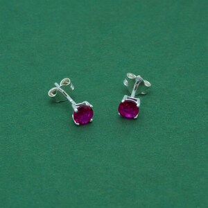 May include: A pair of silver stud earrings featuring oval-cut, magenta-coloured gemstones. The earrings are displayed against a solid green backdrop, highlighting their polished metal and vibrant stones. A classic jewellery item.